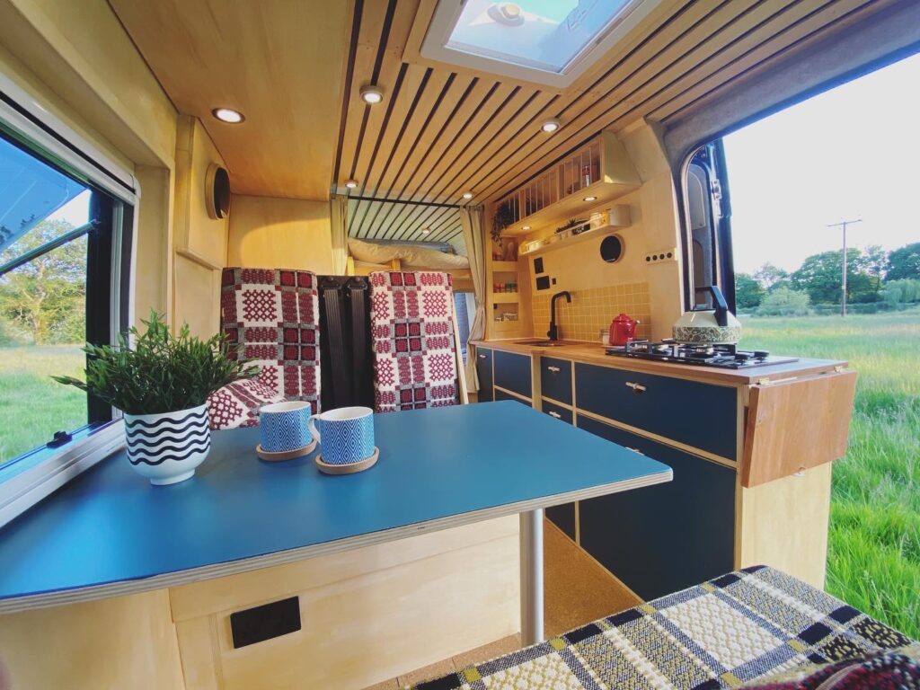 Interior of a well-lit camper van showing a table with potted plants and two mugs, a kitchenette with a stove and cabinets, patterned seating, wood-paneled walls, and a skylight. Windows offer a view of a grassy field and trees outside. The ambiance is cozy and inviting.