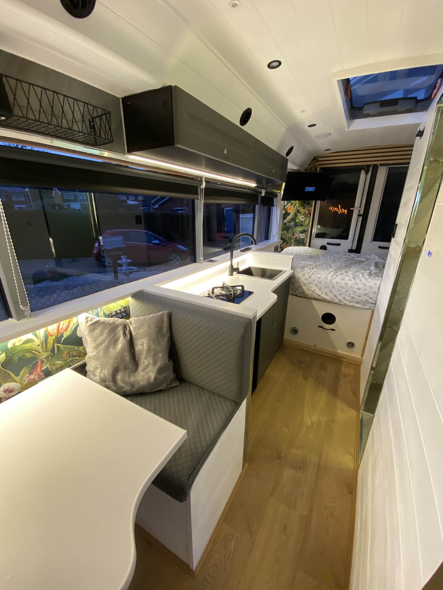 A well-lit, modern camper van interior features a small kitchen with a sink, faucet, and counter space on the left. Adjacent is a cushioned seating area with a table. The back has a cozy bed with polka dot bedding. Light wood flooring and large windows with greenery outside complete the space.