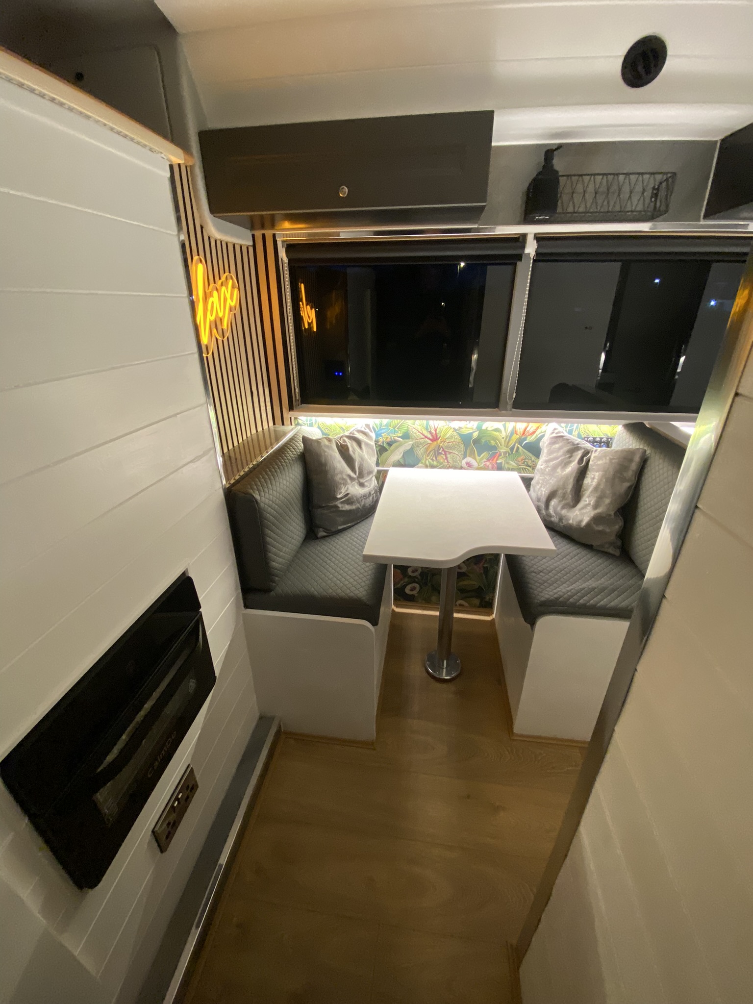 A cozy RV interior featuring a small dining area with two cushioned bench seats and a white table in between. The back wall has colorful, floral wallpaper and a neon sign. The wall on the left is white with a black heater, while the right side has large windows with black shades.