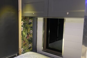 A compact bedroom interior of a van with a cushioned bed in the foreground. The bed faces a sleek, modern window framed with horizontal white wooden panels and dark trim. Above the window are two gray overhead cabinets. The wall to the left features vibrant wallpaper with a floral and bird pattern.