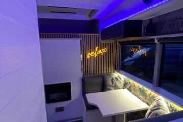 Interior of a modern camper van lit with blue LED lights. A small dining area is visible with two cushioned benches and a white table in between. The wall behind the dining area features a neon sign that reads “relax.” Wood paneling and large windows contribute to the contemporary design.