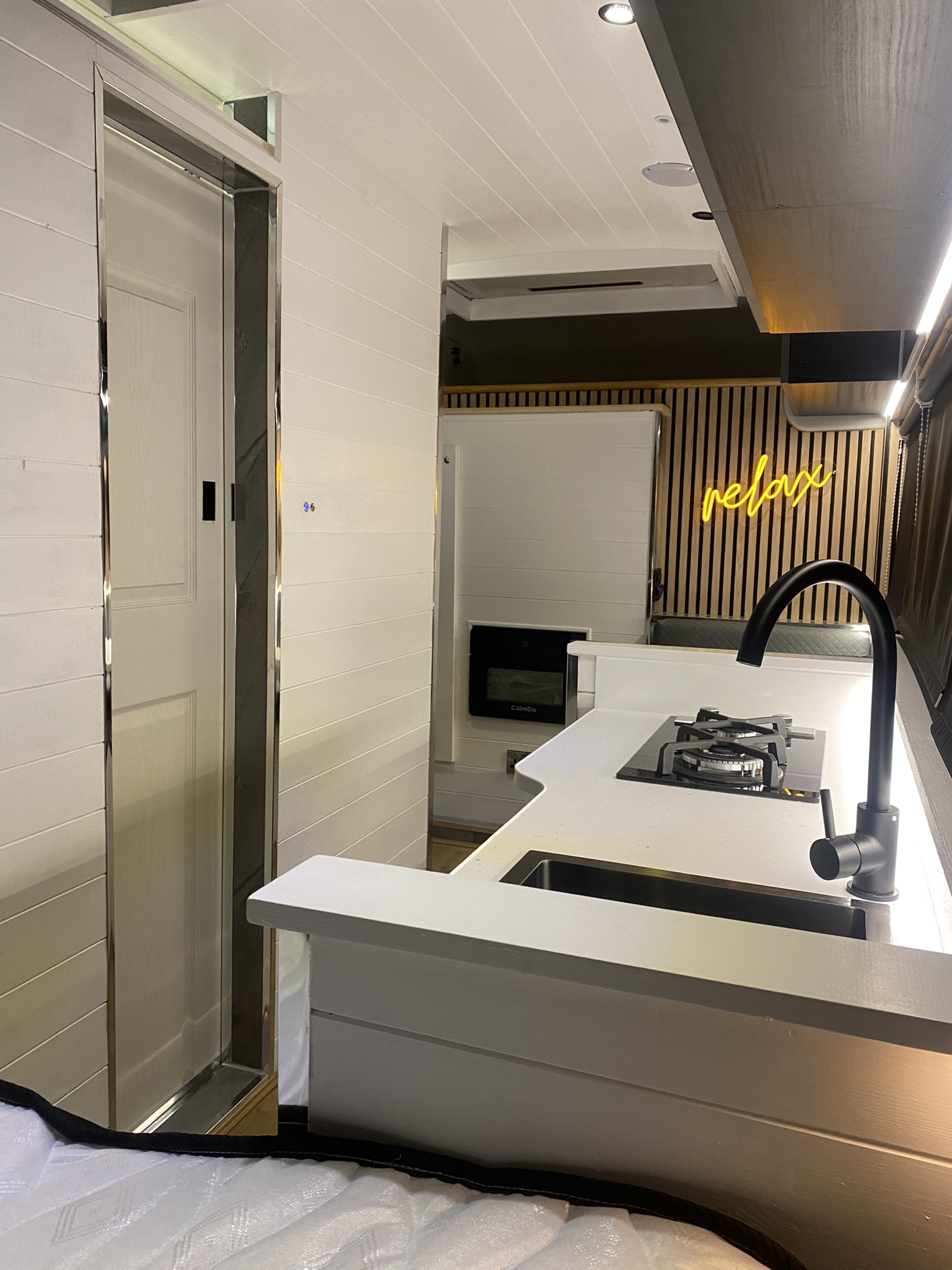 A modern RV interior features sleek white cabinetry and walls. At the center is a compact kitchen with a gas stove, sink, and black faucet. A neon sign reading 