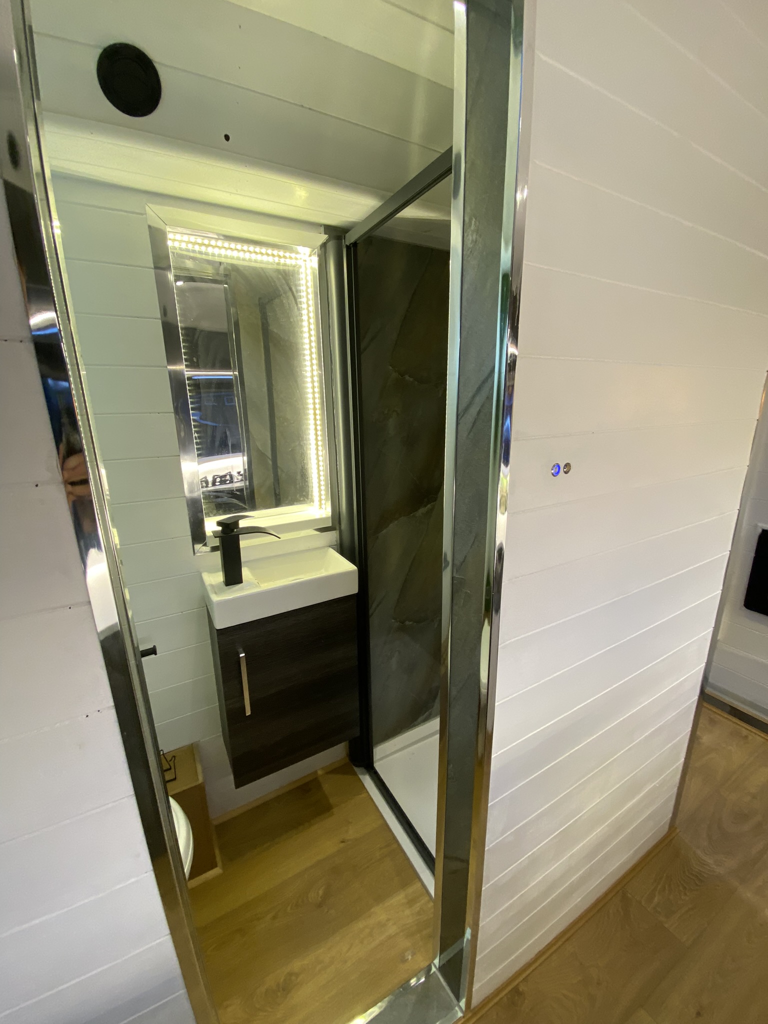 A compact bathroom with a modern design. It features a small black vanity with a white sink and contemporary faucet. Beside the vanity is a shower stall with dark tiles. The space is illuminated by LED strip lighting around the mirror. The walls are white, and the floor is wooden.