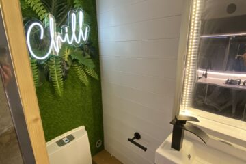 A modern bathroom features a white toilet next to a sink with a black faucet and an illuminated mirror. The left wall is decorated with green artificial plants and a neon sign saying "Chill." The walls have white horizontal paneling, creating a bright and fresh atmosphere.