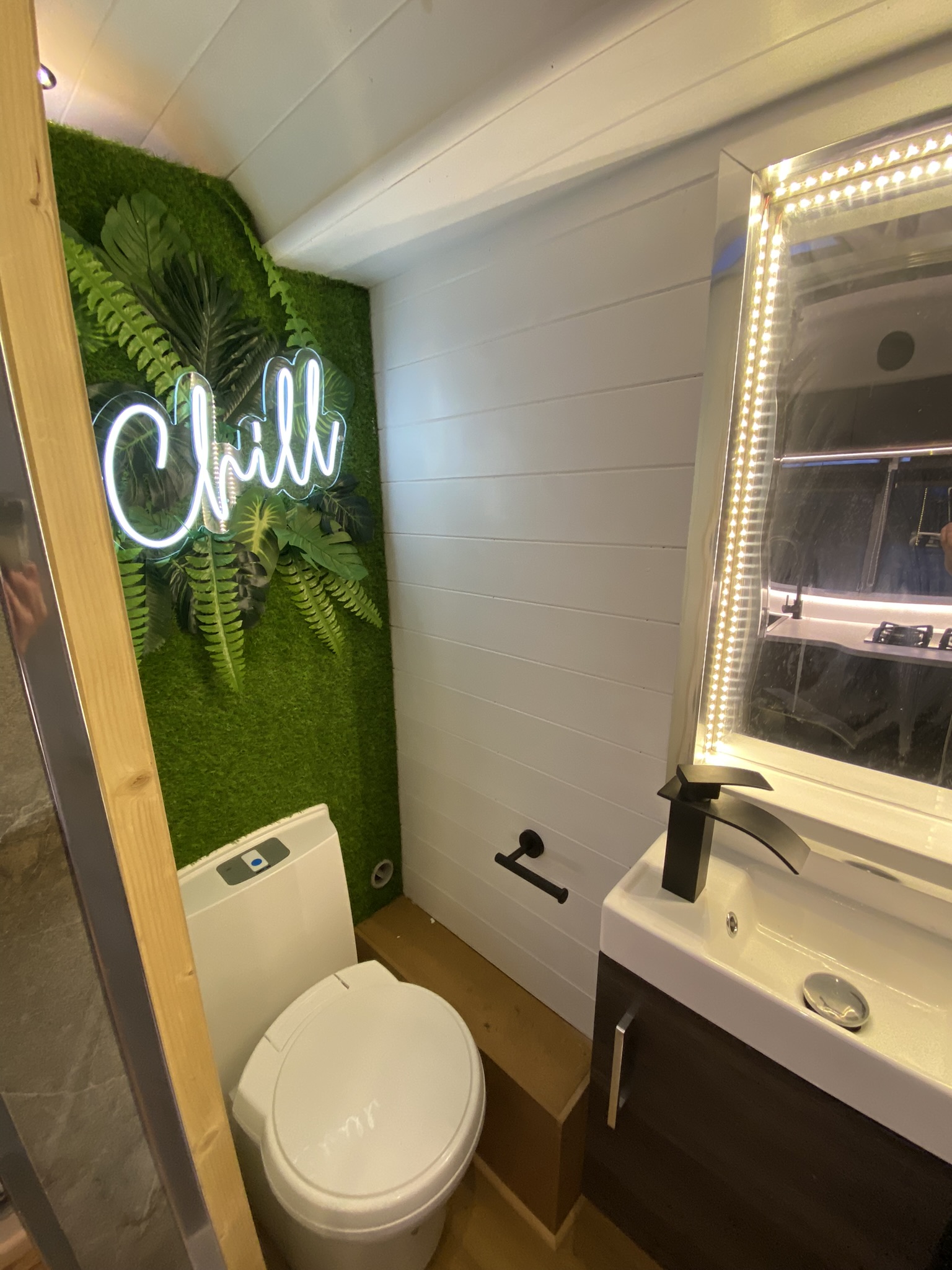 A modern bathroom features a white toilet next to a sink with a black faucet and an illuminated mirror. The left wall is decorated with green artificial plants and a neon sign saying 