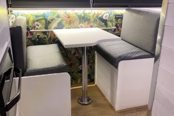 A cozy dining nook in an RV or tiny home, featuring a small square table with two quilted gray booth seats on either side. The walls are adorned with vibrant floral and tropical leaf wallpaper. The windows are covered with closed black blinds, and the floor is a light brown wood.