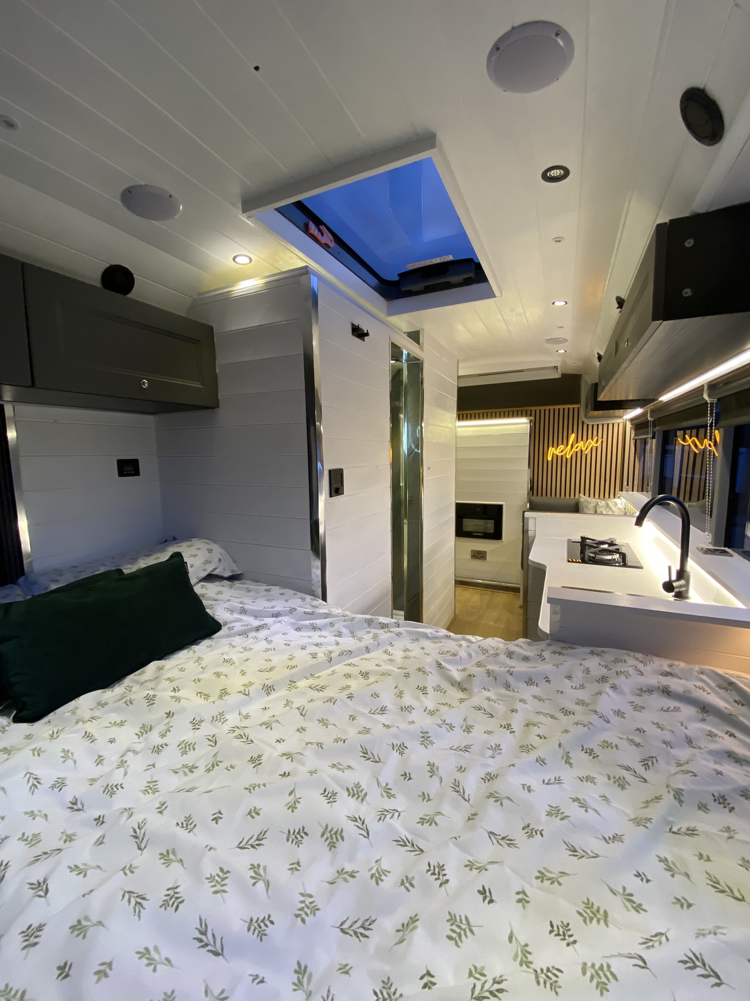 The image shows the interior of a modern, well-lit camper van. There is a bed with a white and green leaf-patterned duvet on the left, a small kitchen with a sink and stove on the right, and a vertical storage cabinet in the back. An open skylight allows natural light to enter.