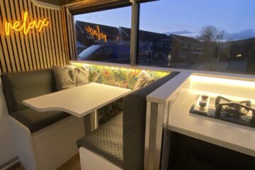 The interior of a modern camper van features a cozy dining area with a white table and two bench seats upholstered in grey quilted fabric. A neon "relax" sign is mounted on the wooden slatted wall. Adjacent is a compact kitchenette with a gas stove, all softly lit by ambient lighting.