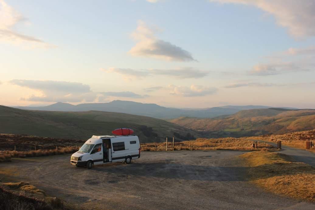 A white camper van with an open door is parked on a gravel clearing. A red kayak is strapped on top of the van. Rolling hills and mountains are seen in the distance under a sky scattered with clouds during a sunset, casting a warm light over the landscape.