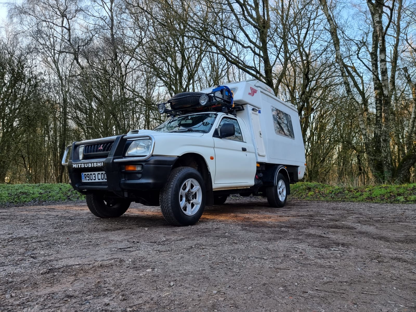 A white 4x4 camper truck is parked on a dirt clearing surrounded by tall, leafless trees. The vehicle features large off-road tires, a roof rack loaded with gear, and a compact camper unit mounted on the back for overlanding. The sky is partly cloudy, suggesting a cool, early spring day.