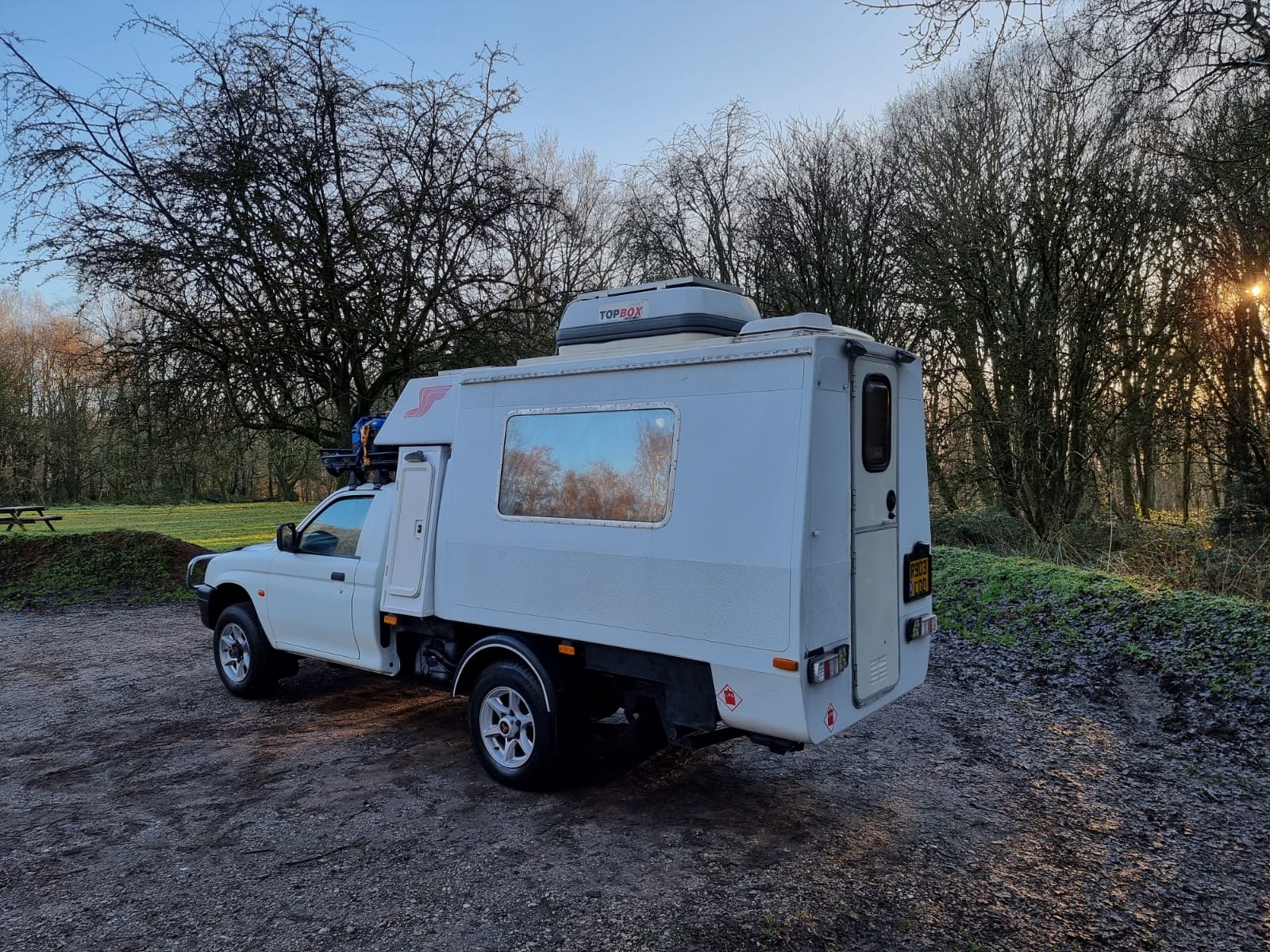 A white camper van is parked on a gravel area amidst a wooded landscape with bare trees. The van has a rectangular structure attached to its back, featuring a roof storage unit and a window. The sky above is clear with hints of the setting or rising sun casting a soft light.