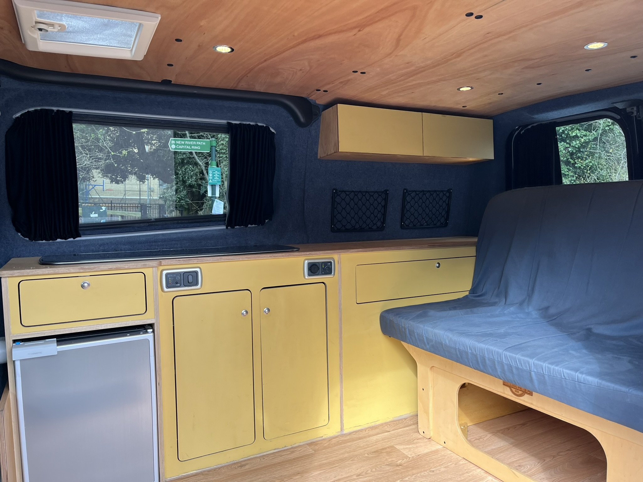 The image shows the interior of a camper van. It features yellow cabinets, a small built-in refrigerator, and a gray cushioned bench. The walls and ceiling are adorned in neutral tones with recessed lighting. A window with blackout curtains is visible, revealing outdoor greenery.