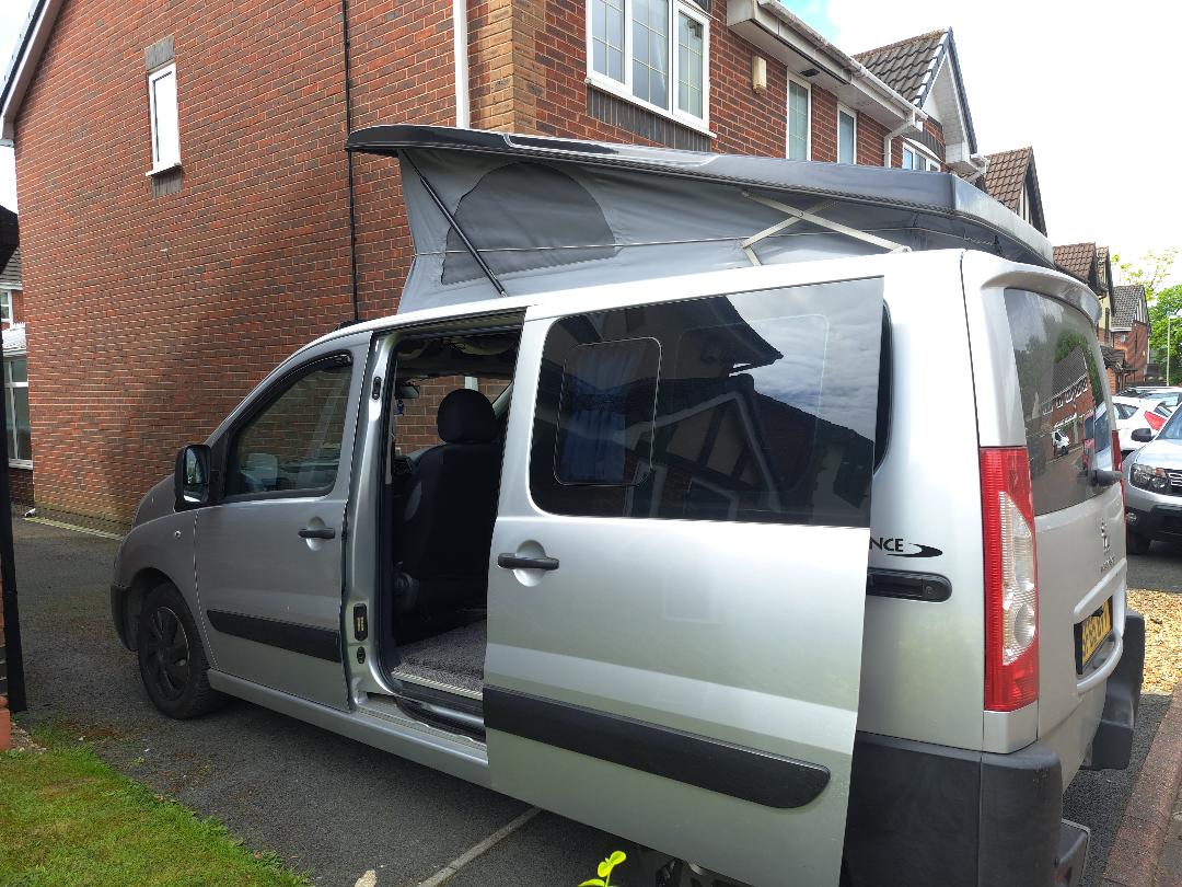 A silver campervan with its side door open and roof extended is parked in front of a brick house. The van has a black interior with the driver's seat visible. The roof is elevated to create additional headroom. The house has white-framed windows and a partially visible second story.