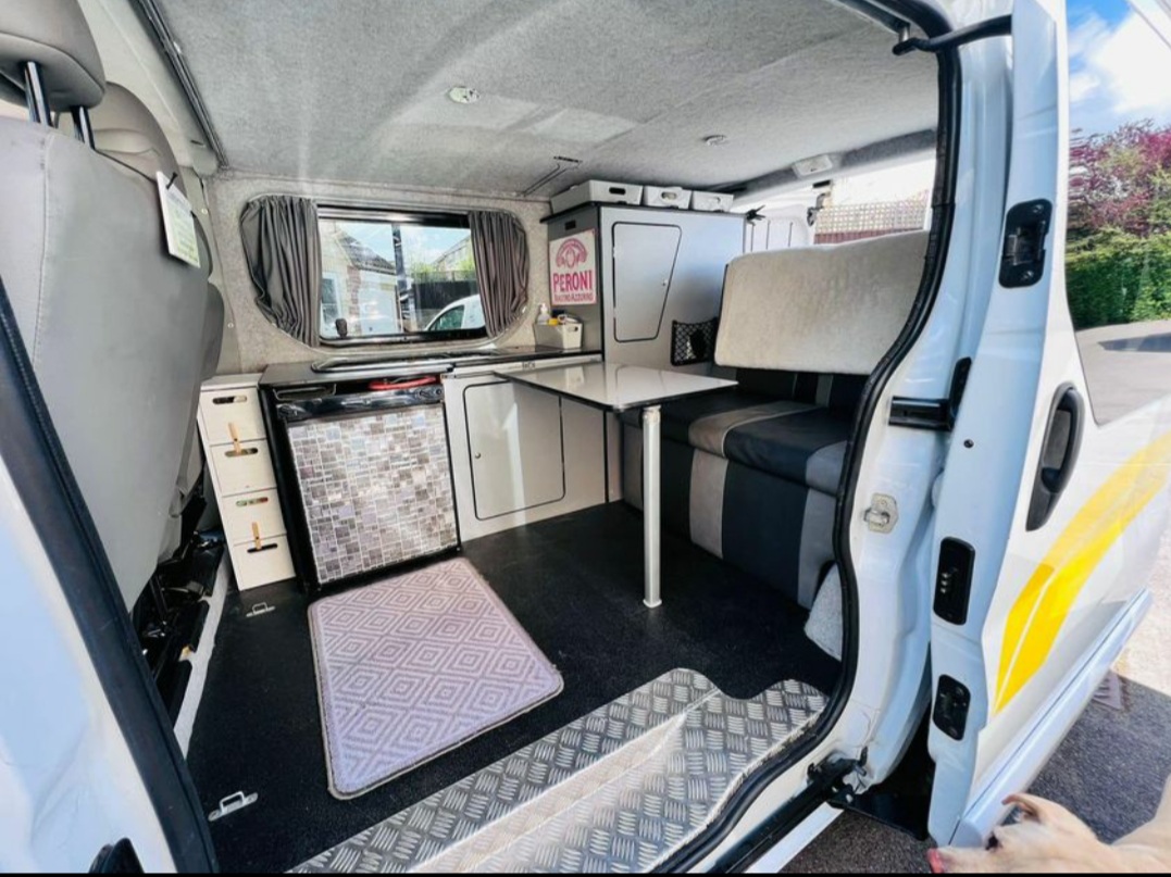 The image shows the interior of a compact, neatly organized camper van. It includes a small kitchenette with a mini fridge, a counter with cabinets, a foldable table, and a cozy bench seat that likely converts into a bed. The van has curtained windows and various storage compartments.