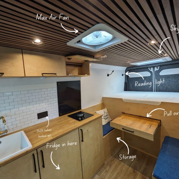 Interior of a camper van featuring light wooden cabinetry and a slatted wooden ceiling. Notable features include labeled Max Air Fan on the ceiling, skylight, reading light above a cozy seating area with a pull-out table, kitchen area with a sink and designated fridge space, and additional storage.