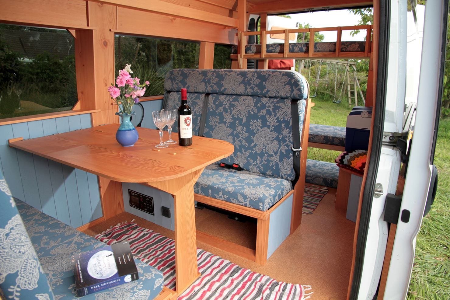 The interior of a cozy camper van with wooden furnishings is shown. A table set with two wine glasses, a bottle of wine, and a vase of pink flowers is at the center. Blue cushioned seats with floral patterns, a striped rug, and a shelf with a bed in the background are also visible.