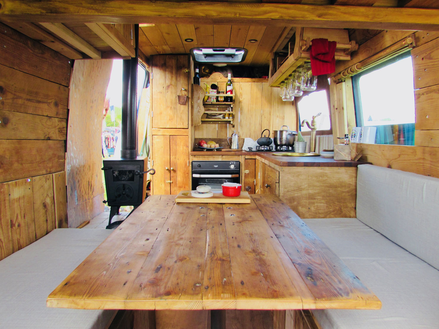 The interior of a rustic wooden camper van includes a central wooden table with benches on either side, a kitchen area with a stove, sink, and shelves filled with kitchenware. A small wood-burning stove is on the left, and a window on the right allows natural light to illuminate the space.