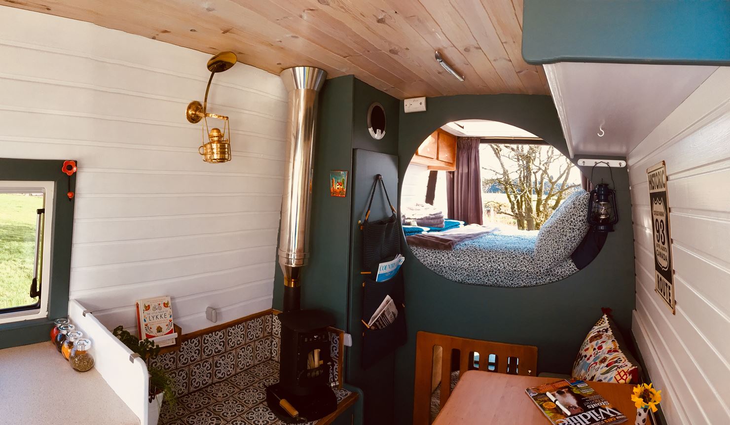 Interior of a cozy camper van featuring a daybed with a round window view, a small kitchen with a sink, a wooden countertop with a colorful pillow and books on it, a wall-mounted lamp, a stove with a rustic chimney, and a wooden ceiling. A potted plant and decorative items add charm.