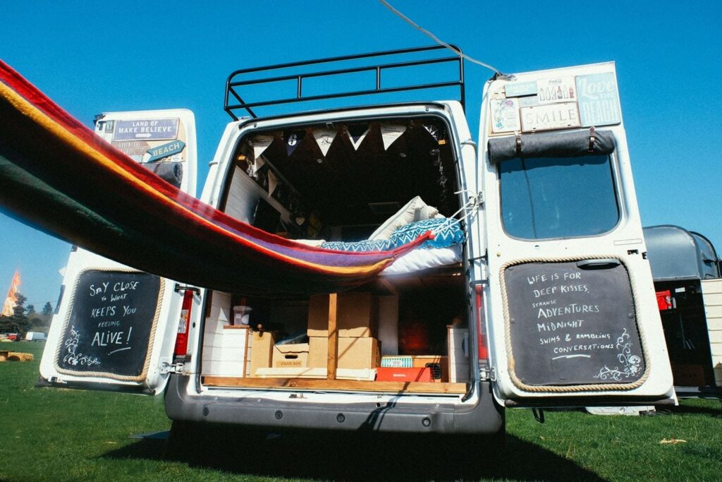 A van with its rear doors open, revealing a cozy interior decorated with colorful fabrics and fairy lights. Handwritten chalkboard signs on the doors display positive messages. A hammock extends from the van, suggesting a relaxed atmosphere. A clear, blue sky and grass surround the scene.