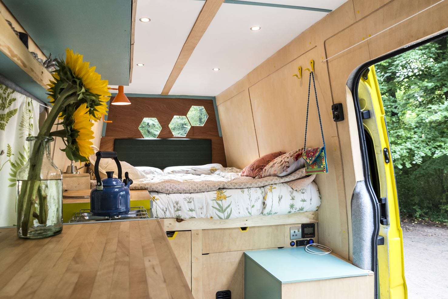 A cozy van interior with wooden walls and ceiling features a neatly made bed with floral bedding and decorative pillows. Hexagonal windows are visible near the head of the bed. Below the bed is storage space. The kitchenette has a wooden countertop, a vase with sunflowers, and a blue kettle.
