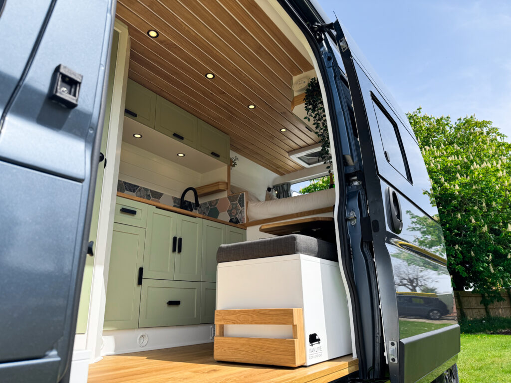 A side view of a camper van with the sliding door open, revealing a cozy, modern interior. The van has wooden ceilings with recessed lights, light green cabinetry, a small sink, a stove, and a seating area with a cushion and storage below. Outside, a grassy area and trees are visible.