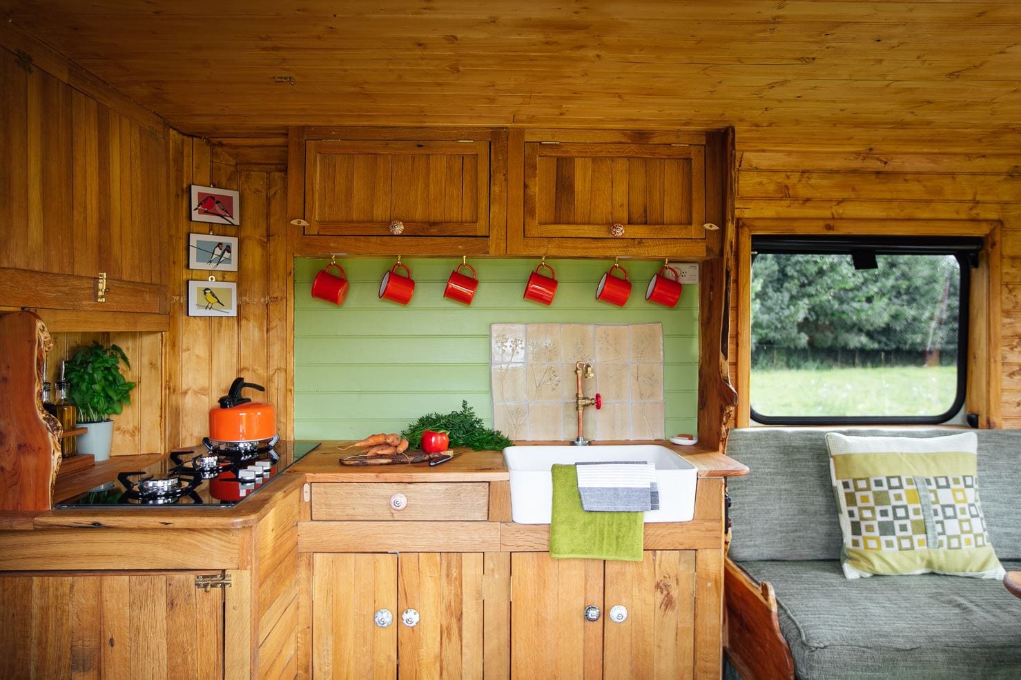 The image shows the interior of a wooden campervan kitchen featuring a countertop with a ceramic sink. Above, there are several red mugs hanging on hooks. To the left, a stove with a pot and several potted plants. To the right, a cushioned seating area with a green throw blanket on it.