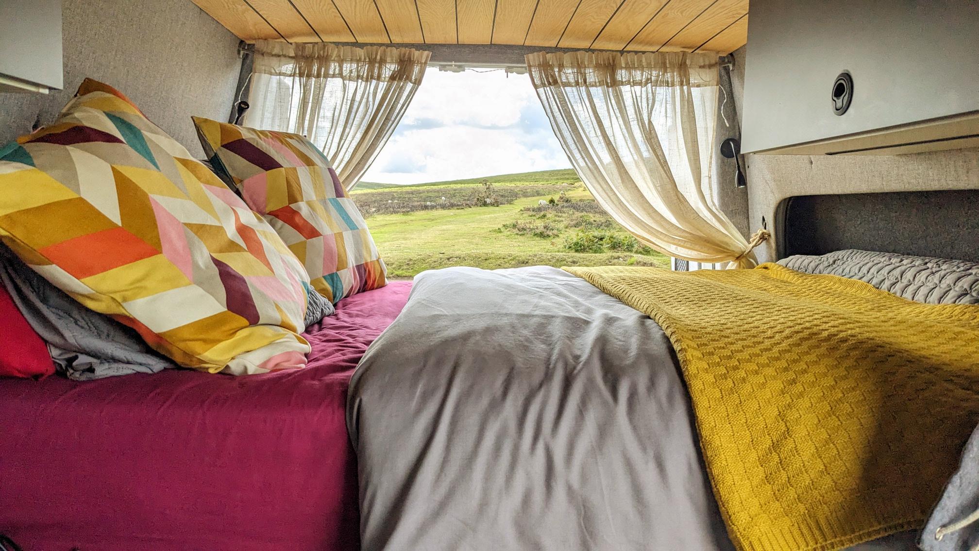 A cozy van bed with vibrant, patterned pillows and a grey duvet is shown. A yellow knit blanket is draped atop the bed. Cream-colored curtains are tied back, revealing a lush, green landscape with a cloudy sky through the van's back door. The wooden ceiling adds warmth to the interior.