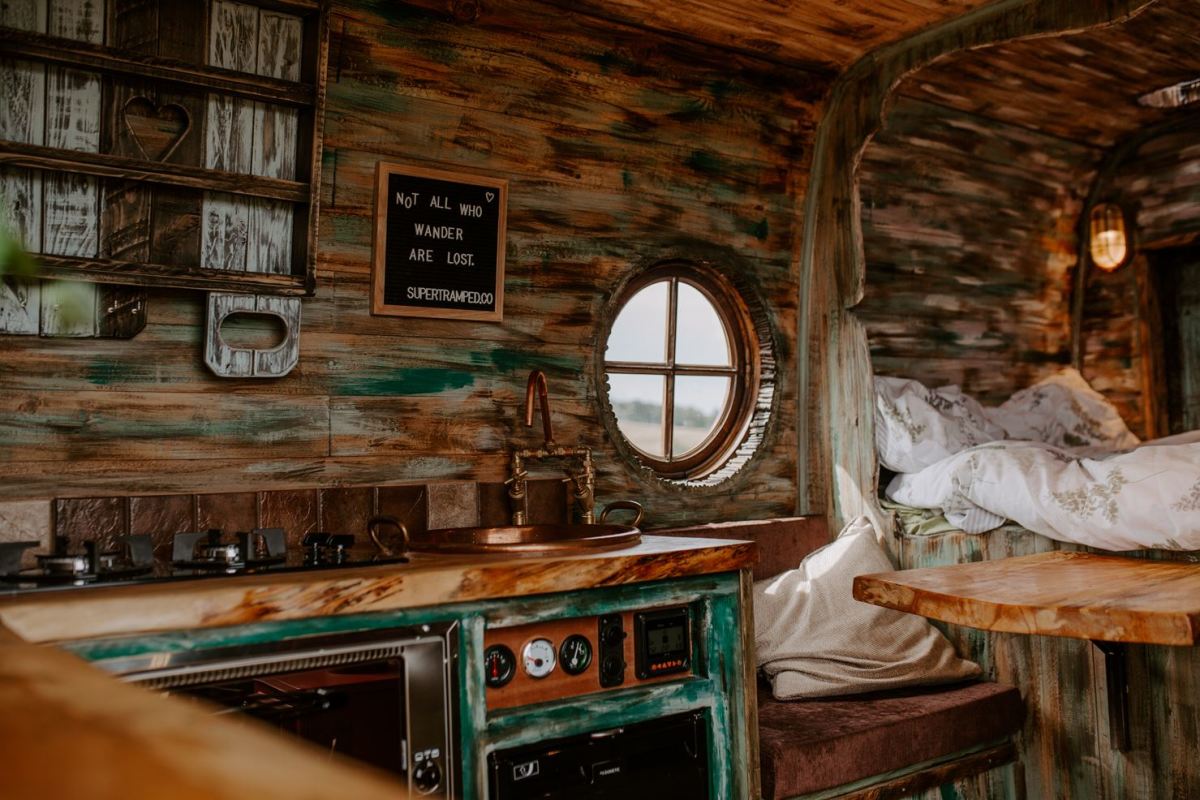 A cozy, rustic camper van interior with a weathered wood finish. The kitchen area features a stove, sink, and open shelving, with a small round window above. Adjacent is a bed with white linens. A sign on the wall reads "NOT ALL WHO WANDER ARE LOST.