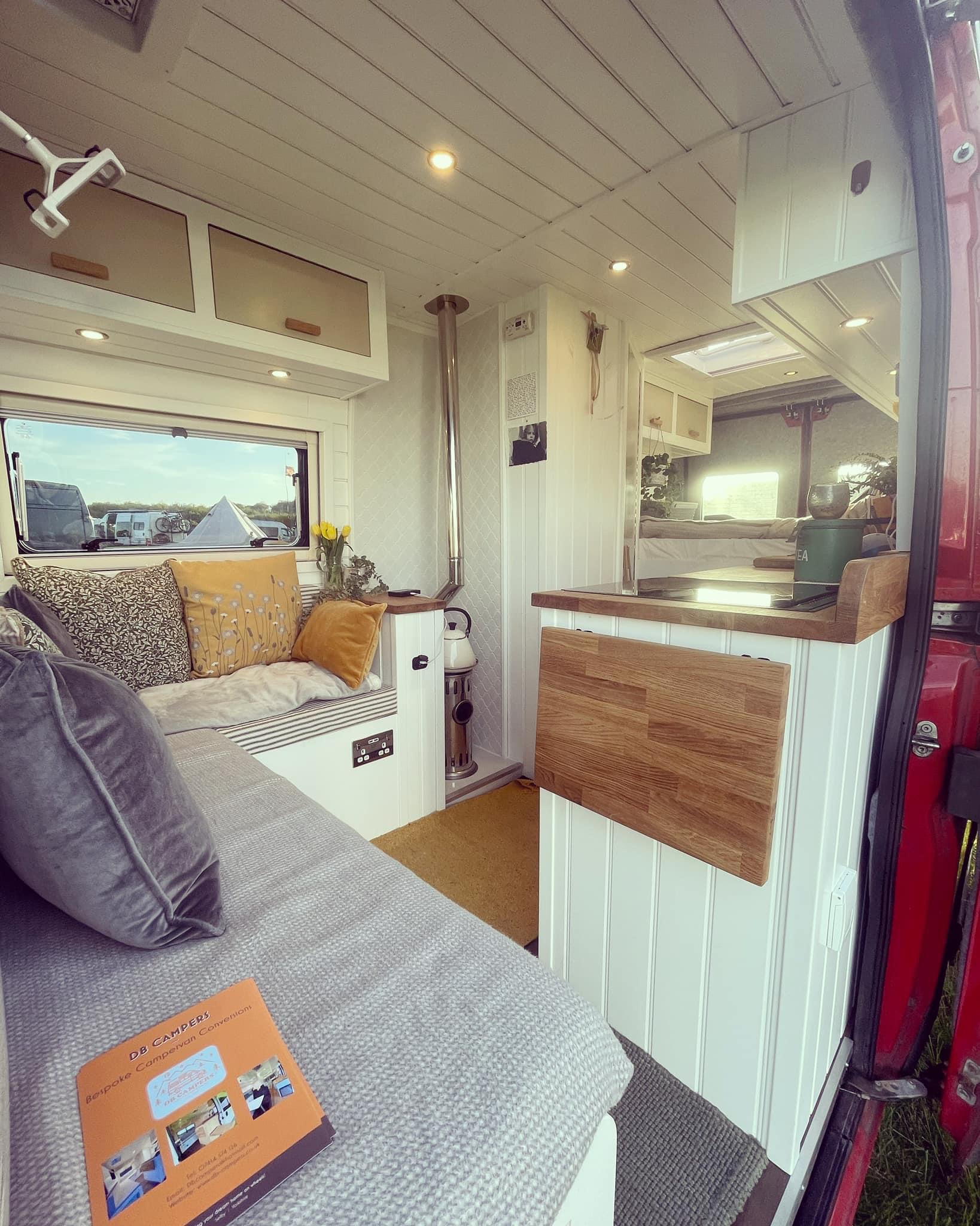 A cozy camper van interior with wooden countertops and white paneling. The seating area features patterned and gray cushions beside a window. A small wood stove and kitchen sink are visible. A book titled