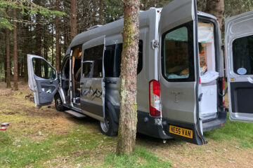 A silver camper van is parked in a forest, with its side and rear doors open. Inside, various camping gear and blankets are visible. Trees surround the vehicle. The van has a customized plate reading "NE55 VAN." Sunlight filters through the leaves, creating a peaceful outdoor setting.