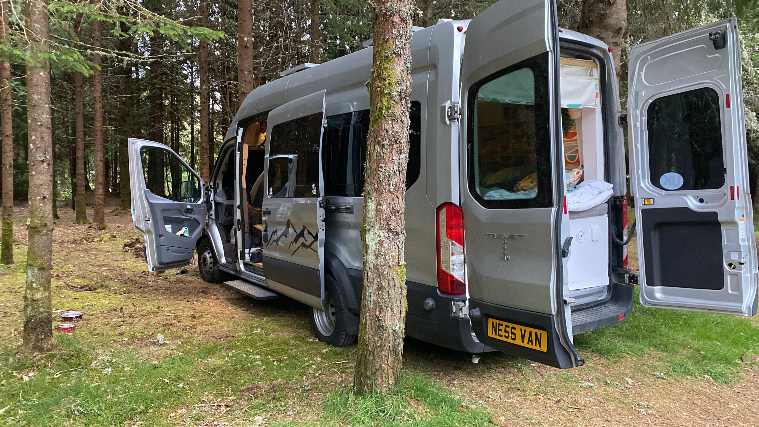 A silver camper van is parked in a forest, with its side and rear doors open. Inside, various camping gear and blankets are visible. Trees surround the vehicle. The van has a customized plate reading 