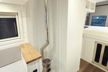 A compact, modern RV interior featuring a small wood stove with a shiny metal chimney pipe extending through the white paneled ceiling. Adjacent to the stove, there is a built-in bench with striped cushions and a wooden countertop. Shelves and storage cabinets are visible in the background.