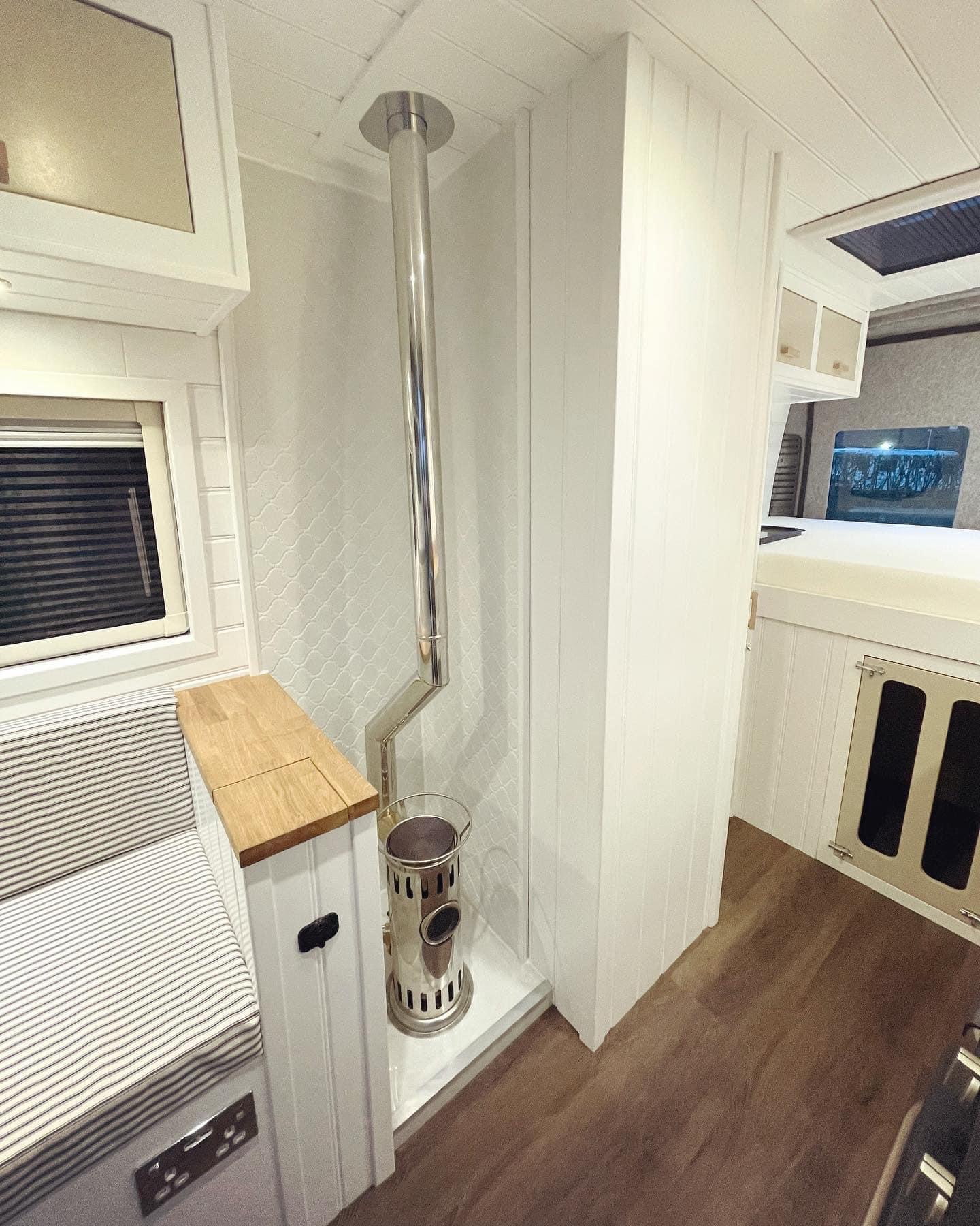 A compact, modern RV interior featuring a small wood stove with a shiny metal chimney pipe extending through the white paneled ceiling. Adjacent to the stove, there is a built-in bench with striped cushions and a wooden countertop. Shelves and storage cabinets are visible in the background.
