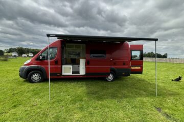 A red camper van parked on a grassy field under a cloudy sky. The side awning is extended. The sliding door is open, revealing the interior. The back doors are closed. A black and white dog lies on the grass near the right rear of the van. Trees and a fence are in the background.