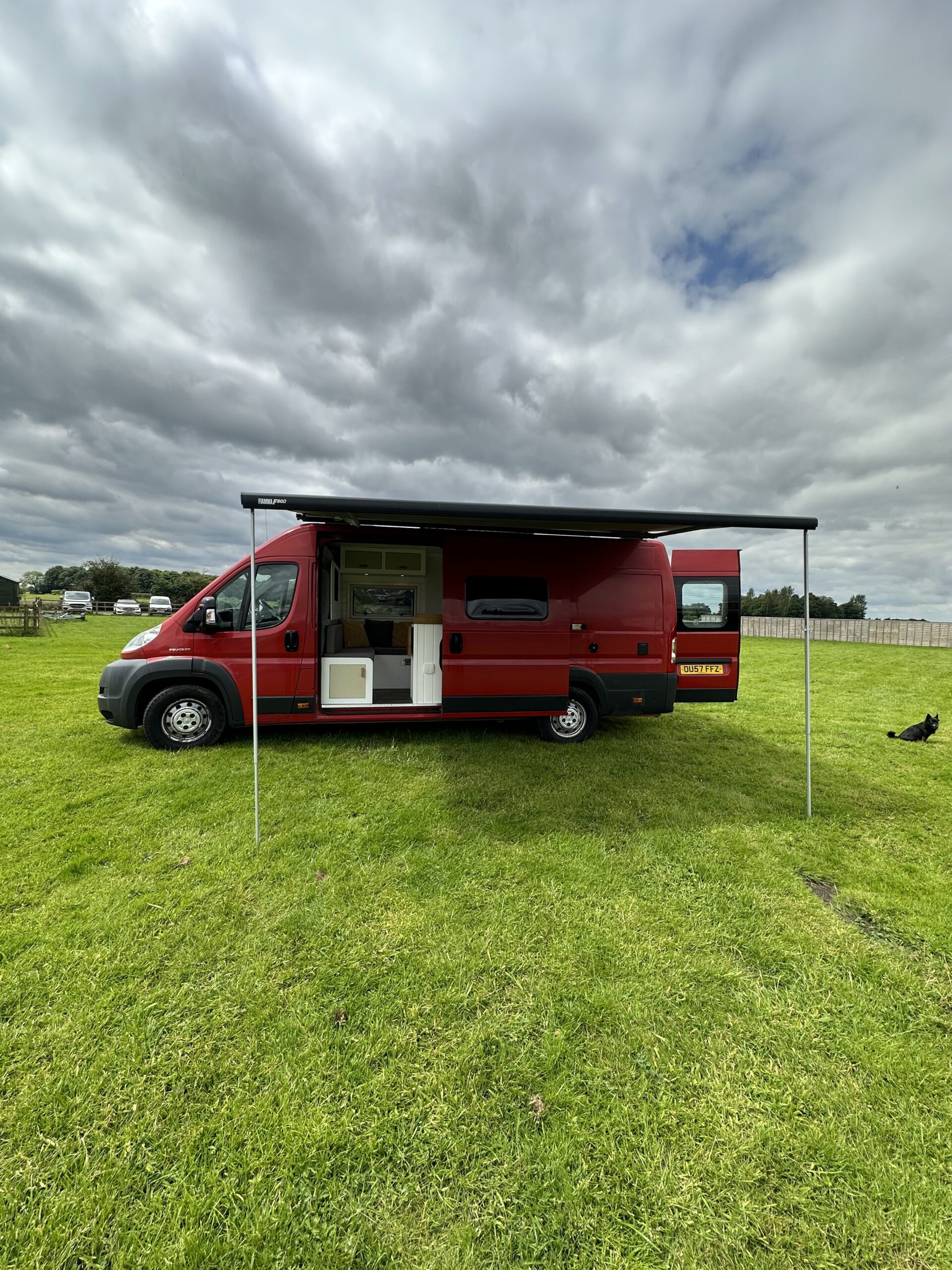 A red camper van parked on a grassy field under a cloudy sky. The side awning is extended. The sliding door is open, revealing the interior. The back doors are closed. A black and white dog lies on the grass near the right rear of the van. Trees and a fence are in the background.