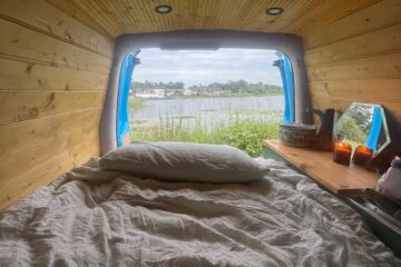 View from inside a cozy camper van with wooden interior walls and ceiling, featuring an unmade bed adorned with patterned linens. A small table on the right holds a basket, a mirror, and lit candles. Through the open back doors, a scenic lakeside landscape is visible under a cloudy sky.