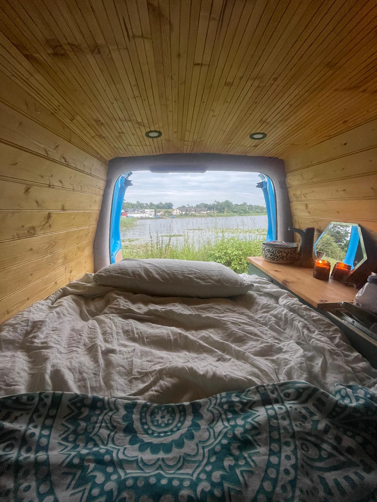 View from inside a cozy camper van with wooden interior walls and ceiling, featuring an unmade bed adorned with patterned linens. A small table on the right holds a basket, a mirror, and lit candles. Through the open back doors, a scenic lakeside landscape is visible under a cloudy sky.