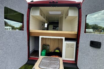 A red van with its back doors open reveals a compact living area. The setup includes a neatly arranged kitchenette with white cabinets, a wooden countertop, and shelves. Below the counter is storage space holding camping gear and a water container. The van is parked on a grassy field.