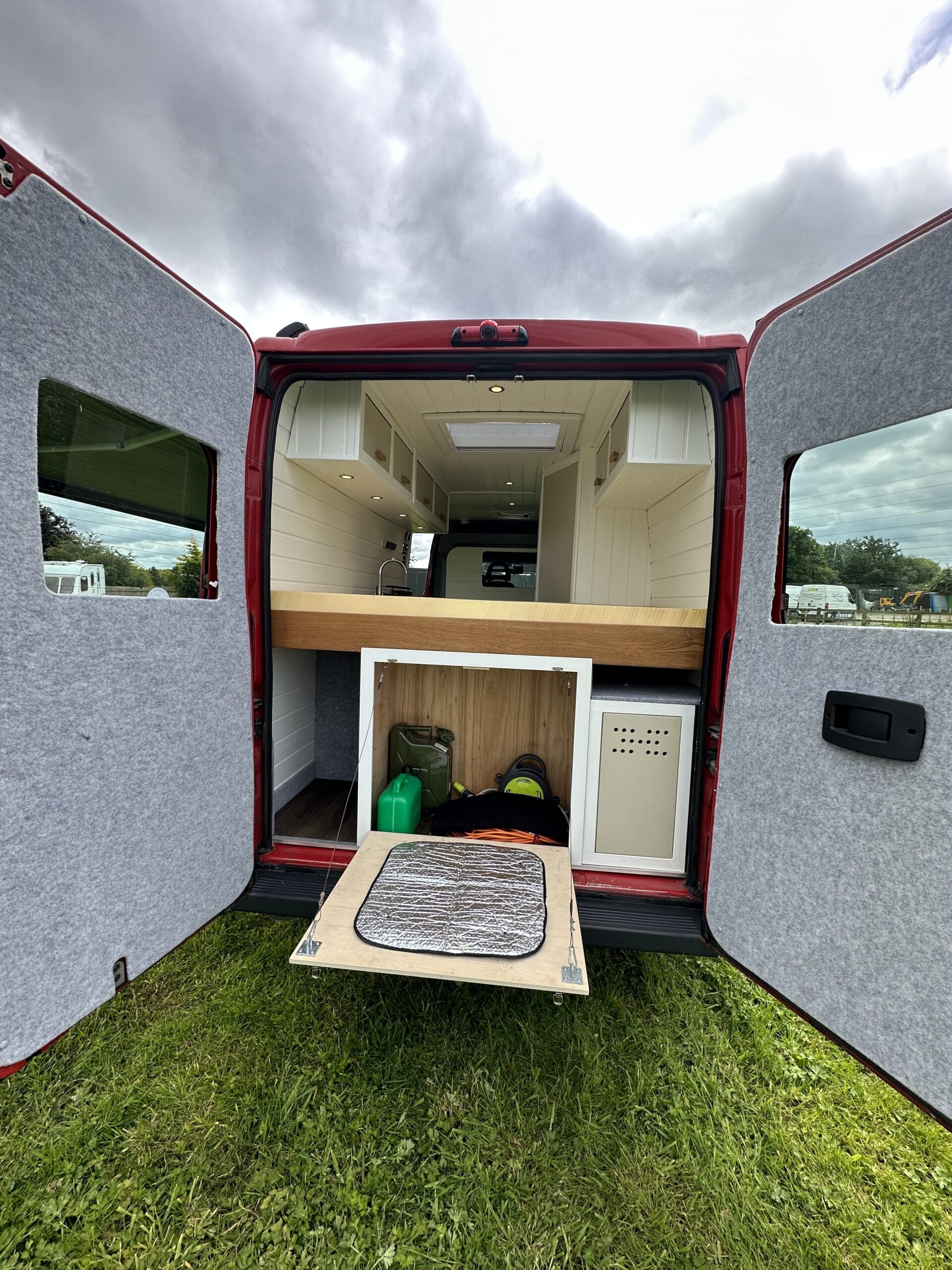 A red van with its back doors open reveals a compact living area. The setup includes a neatly arranged kitchenette with white cabinets, a wooden countertop, and shelves. Below the counter is storage space holding camping gear and a water container. The van is parked on a grassy field.