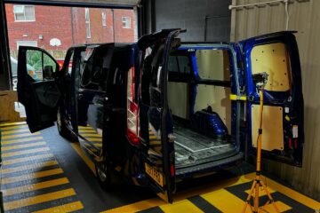 A blue van with its side and rear doors open is undergoing maintenance in a garage. The interior paneling has been removed, exposing the metallic framework. The van is parked on a floor with yellow and black striped markings. A tripod light stand is positioned near the rear of the van.