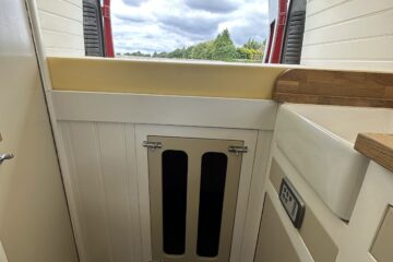 A compact kitchen area in a camper van with white paneled cabinets and wooden accents. A cutout window at the back offers a view of a cloudy sky and greenery. The floor is dark wood, and there's a small control panel on the side wall. The space is bright and neatly organized.
