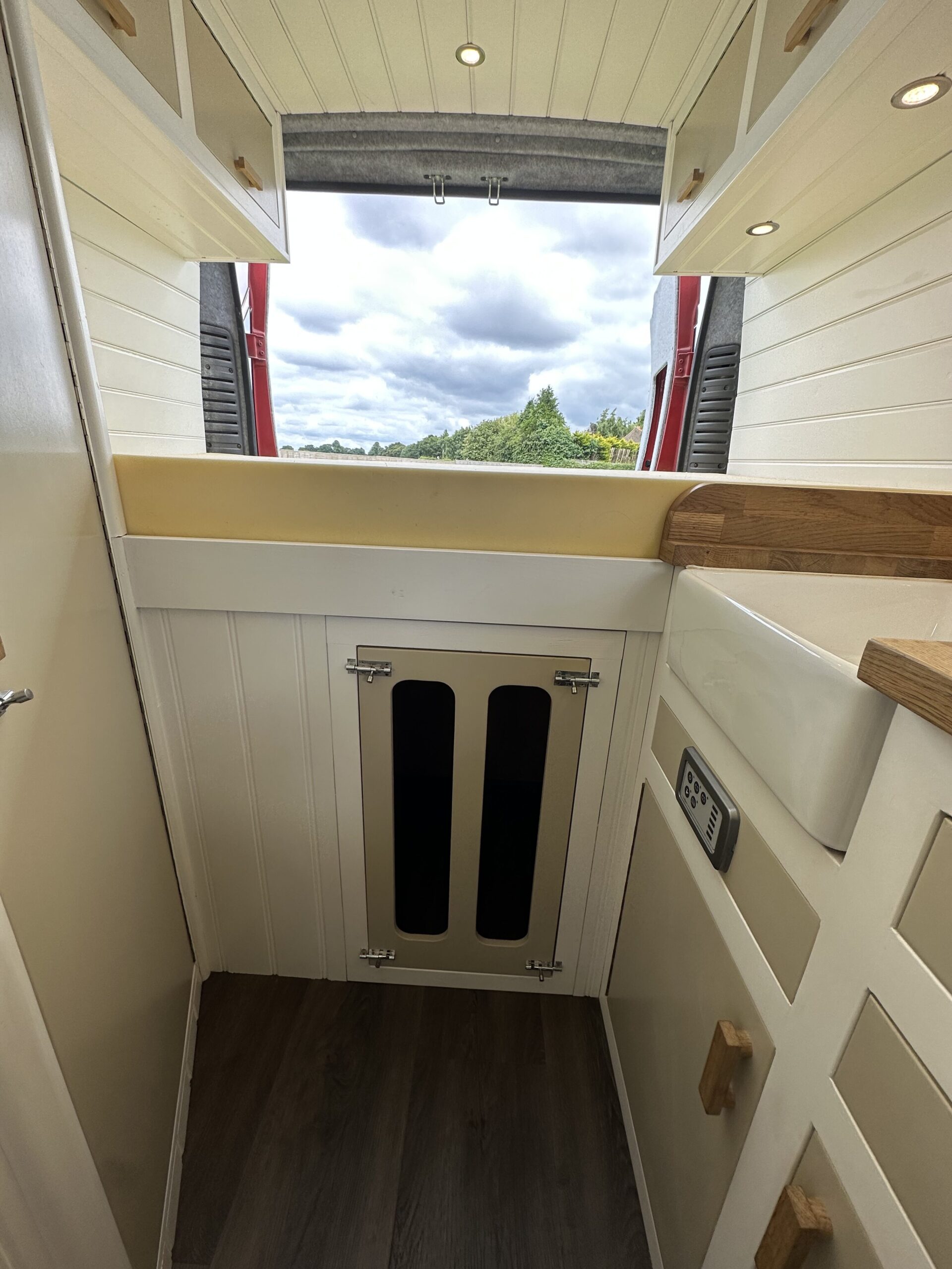 A compact kitchen area in a camper van with white paneled cabinets and wooden accents. A cutout window at the back offers a view of a cloudy sky and greenery. The floor is dark wood, and there's a small control panel on the side wall. The space is bright and neatly organized.