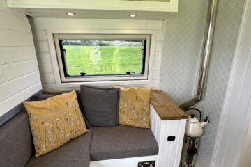 A cozy van interior features a corner seating area with grey cushions and two yellow pillows with white floral patterns. Above, there are two beige cabinet doors. A window shows a green outdoor scene. A wood-burning stove with a metal chimney sits beside the seating area. The floor is dark wood.