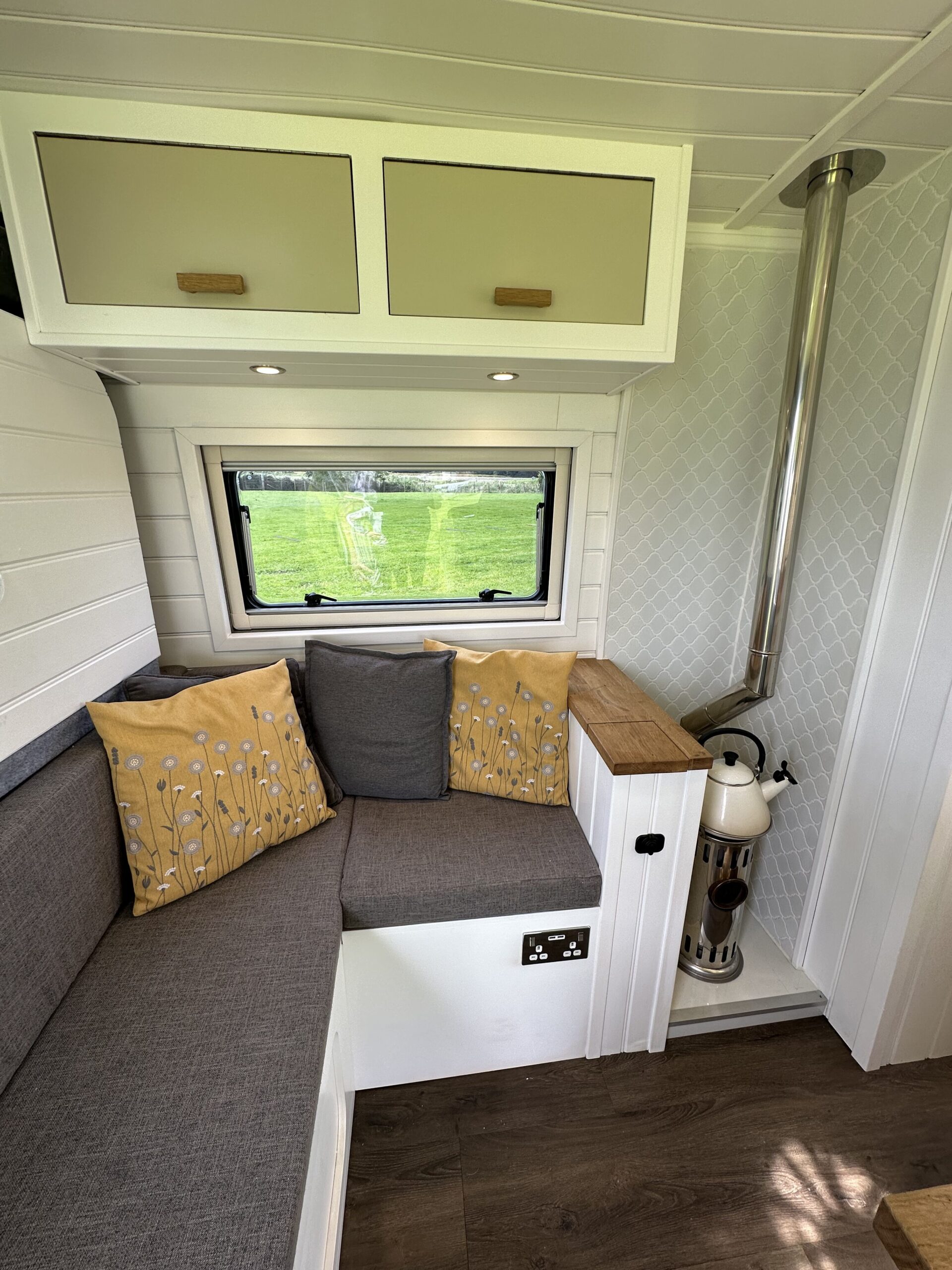 A cozy van interior features a corner seating area with grey cushions and two yellow pillows with white floral patterns. Above, there are two beige cabinet doors. A window shows a green outdoor scene. A wood-burning stove with a metal chimney sits beside the seating area. The floor is dark wood.