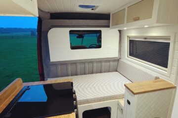 This image shows the interior of a camper van featuring a cozy wooden countertop kitchen area with a built-in stove on one side, and a bed on the other. The bed is positioned below a large window. Cabinets are mounted on the ceiling for storage, and wood and white finishes create a warm and airy atmosphere.