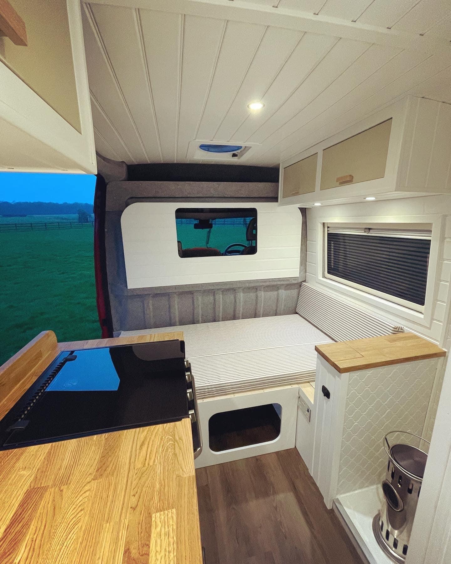 This image shows the interior of a camper van featuring a cozy wooden countertop kitchen area with a built-in stove on one side, and a bed on the other. The bed is positioned below a large window. Cabinets are mounted on the ceiling for storage, and wood and white finishes create a warm and airy atmosphere.