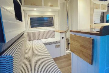 Interior of a modern camper van featuring a long, striped cushioned bench seat along the left wall below a window with closed blinds. Wooden surfaces, white paneling, and recessed lighting create a cosy atmosphere. A foldable table is attached to a wooden divider on the right.