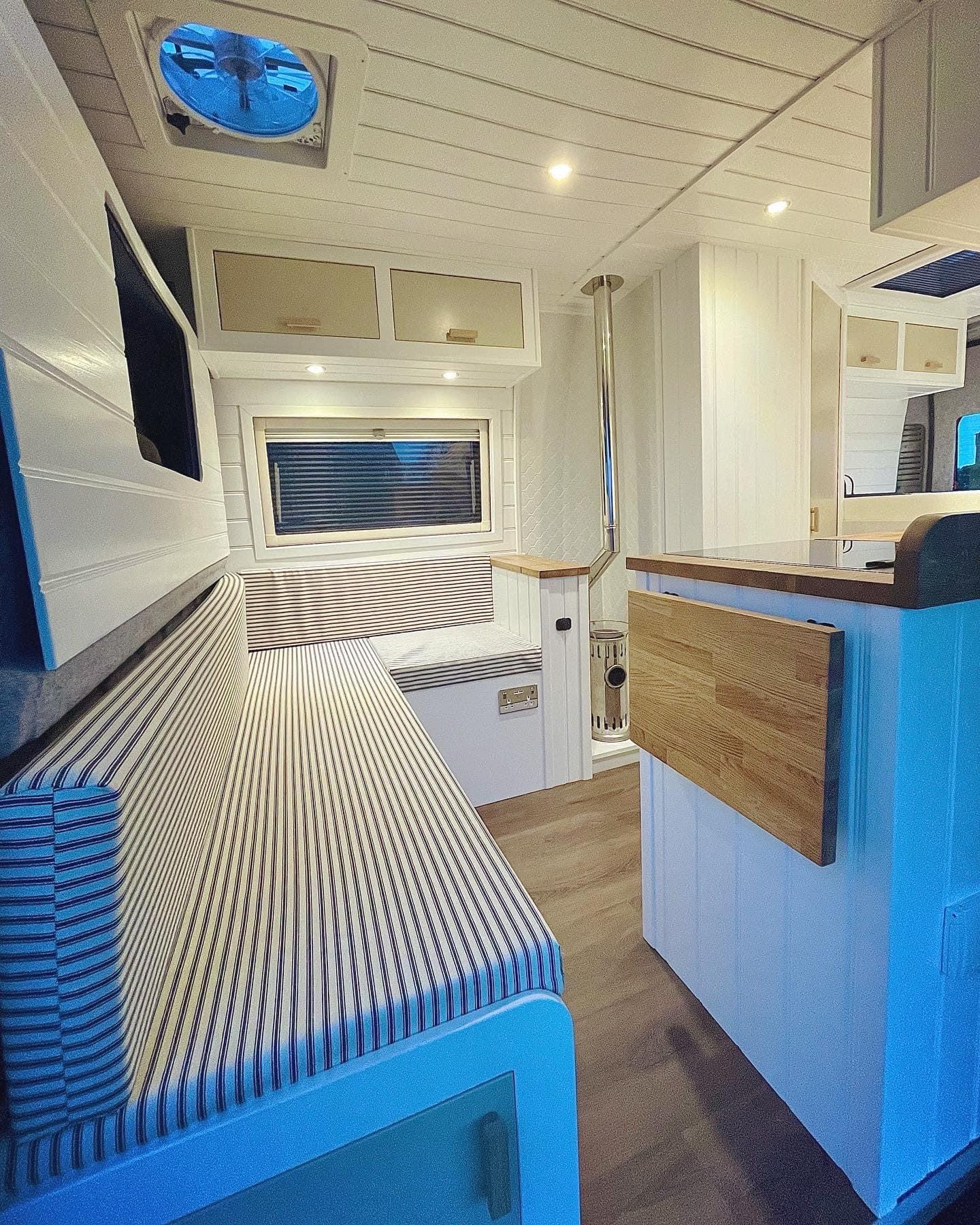 Interior of a modern camper van featuring a long, striped cushioned bench seat along the left wall below a window with closed blinds. Wooden surfaces, white paneling, and recessed lighting create a cosy atmosphere. A foldable table is attached to a wooden divider on the right.