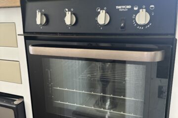 The image shows a Thetford Triplex oven installed in a kitchen setting. The oven has a black control panel with four knobs and markings for temperature and settings. The oven door has a glass window, and there is a vented handle. Light streams in through a nearby window, illuminating the scene.