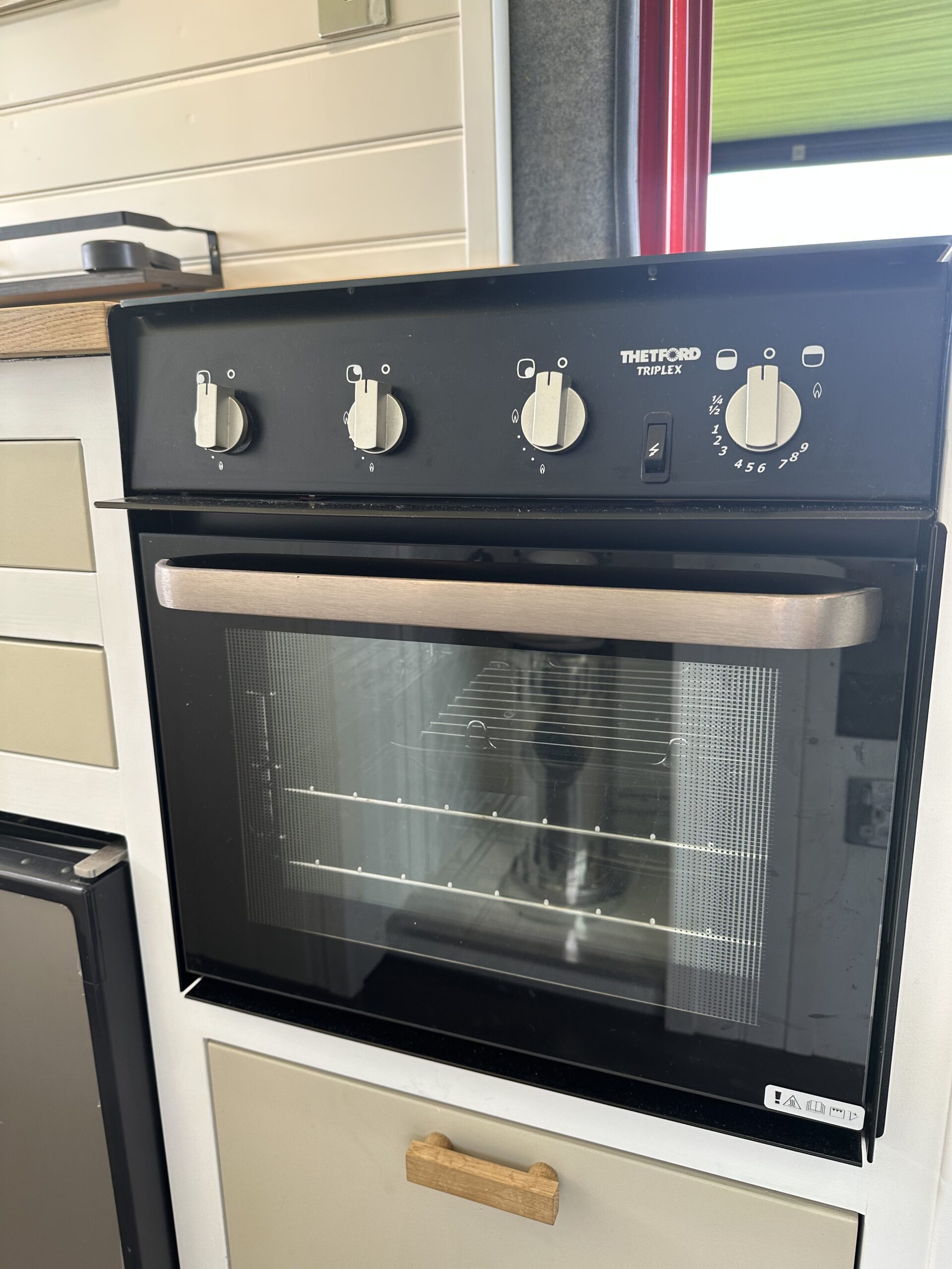 The image shows a Thetford Triplex oven installed in a kitchen setting. The oven has a black control panel with four knobs and markings for temperature and settings. The oven door has a glass window, and there is a vented handle. Light streams in through a nearby window, illuminating the scene.